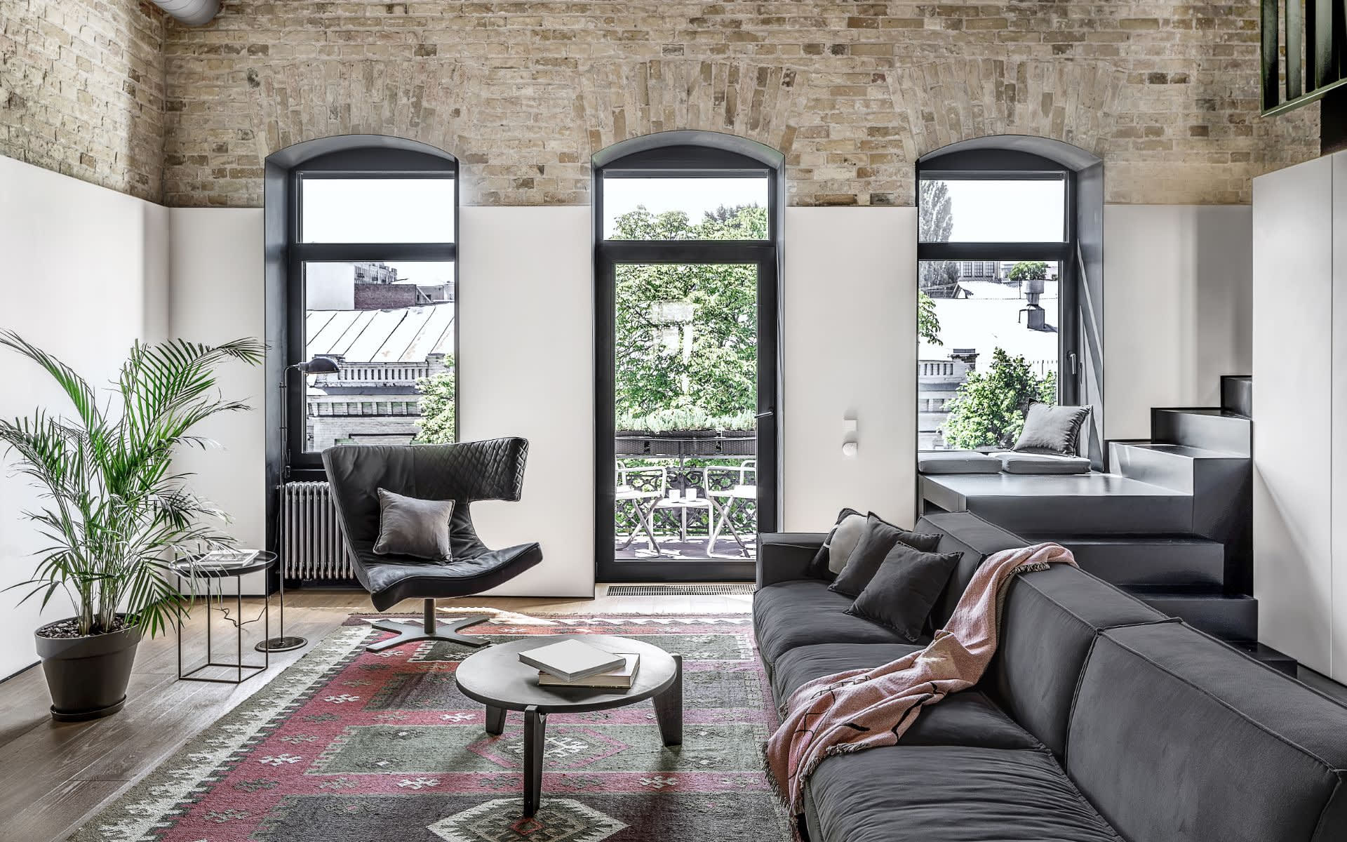 Three large arched windows with black frames offer views of outdoor trees and buildings