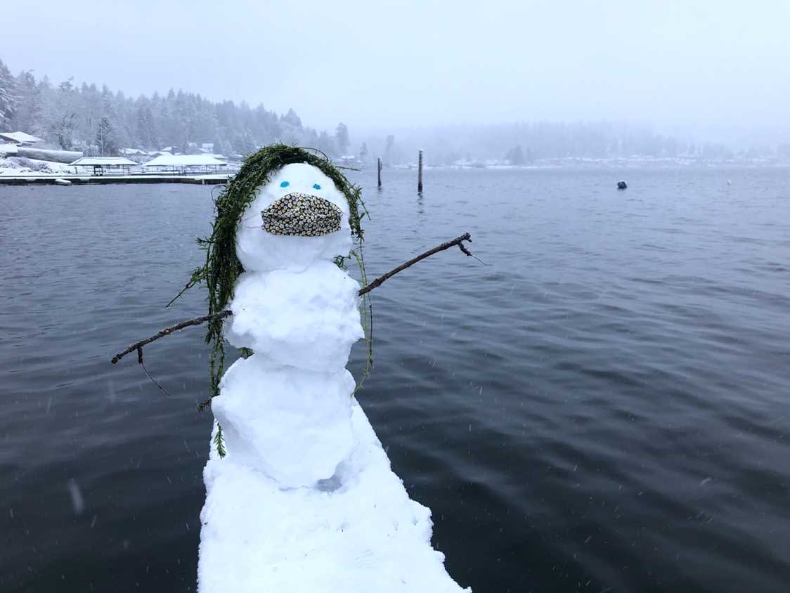 A snowman wearing a face mask and standing on a dock in a lake