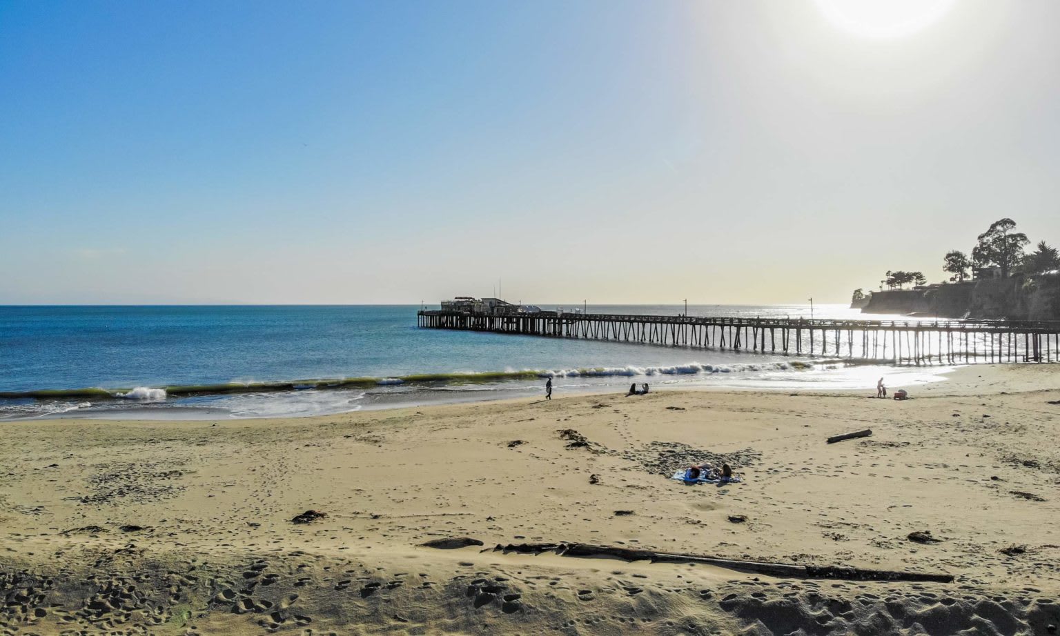 Sandy beach and long wooden pier over blue water, with trees on the headland.