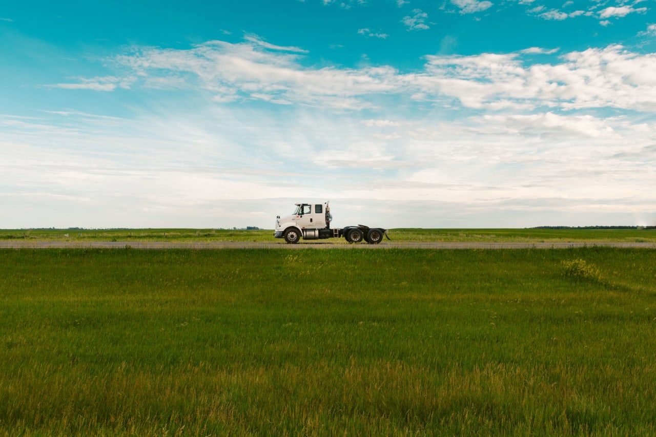 A white semi truck driving on a straight highway with a green field on the side.