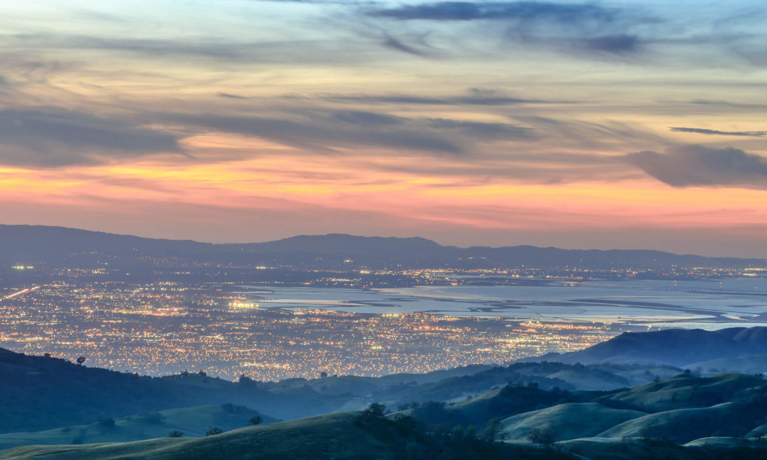City lights and shoreline wetlands seen from rolling hills beneath a colorful dusk sky.