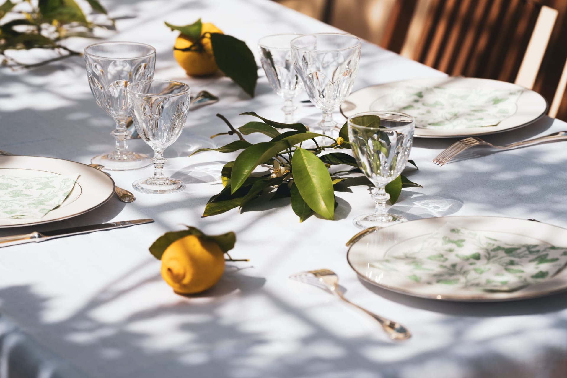 Sunlit table setting with crystal glasses, patterned plates, lemons, and leafy greenery.