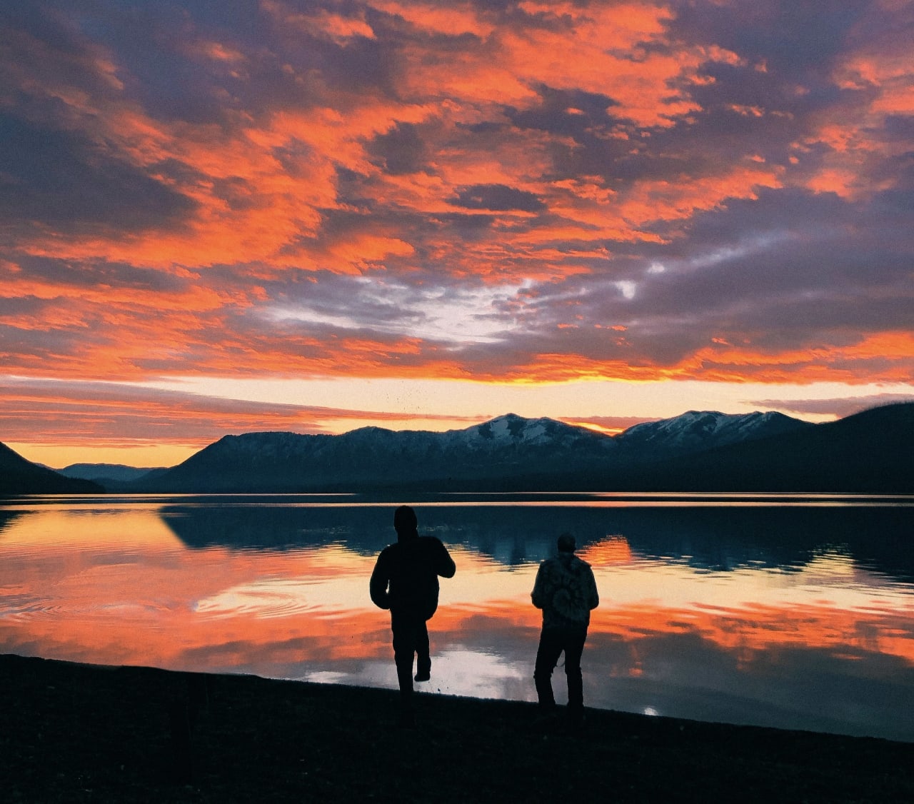 Two people trekking while looking at the colorful sunset over a calm lake.