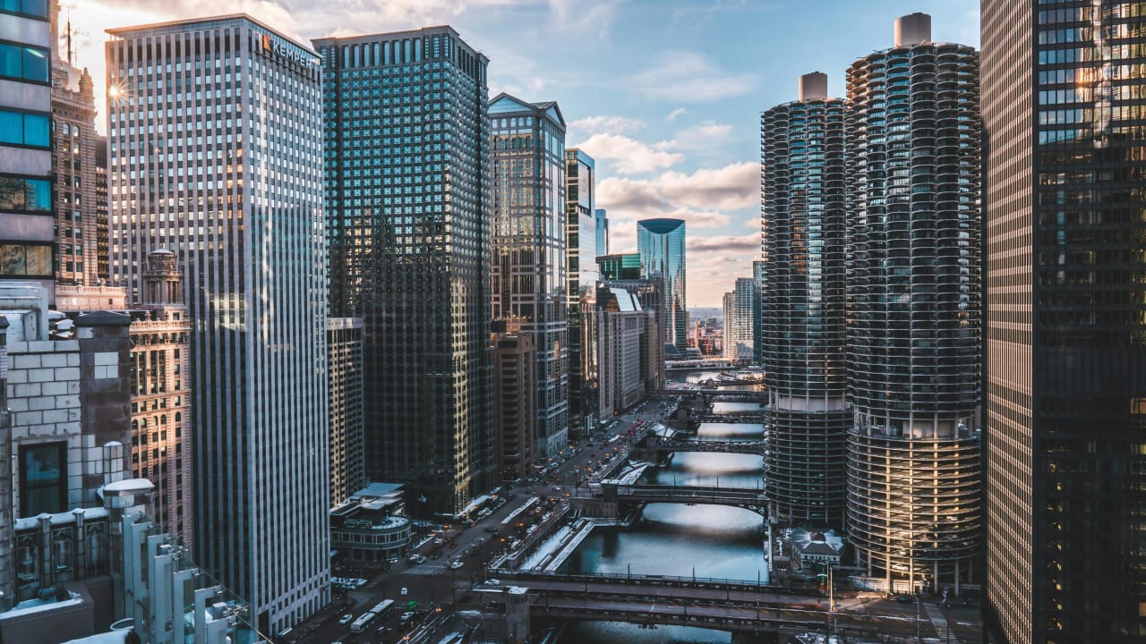 A bustling cityscape with modern glass and metal skyscrapers lining a river, complemented by bridges in the foreground.