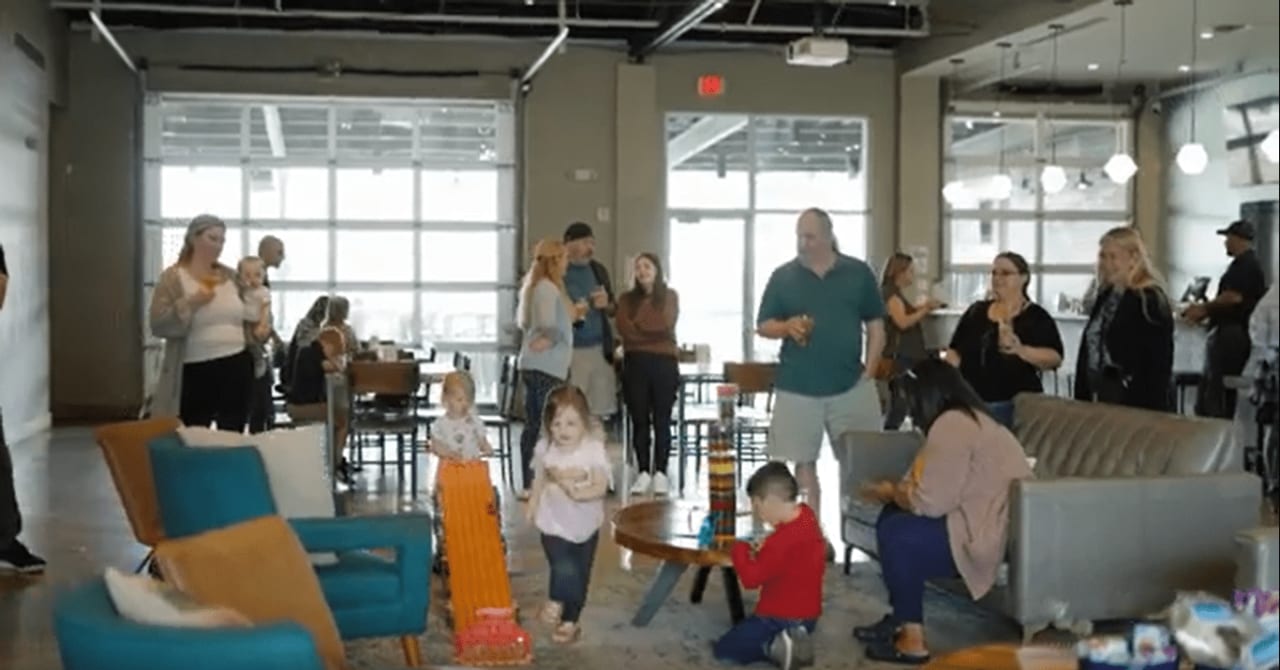 A group of people gathered inside a large room with ample natural light. The setting appears to be a social or community event with participants interacting and moving around.