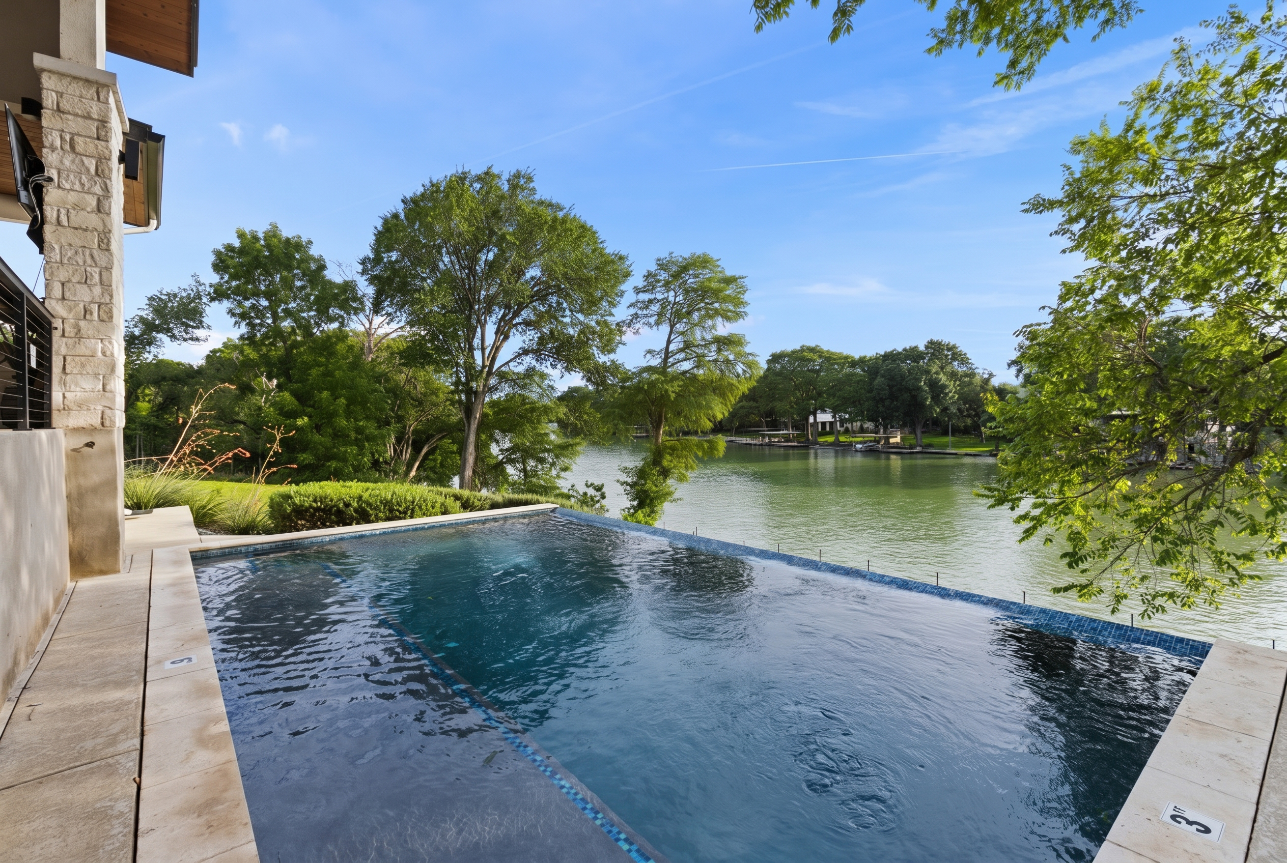 Infinity-edge pool with a blue tile edge and stone coping, looking out across Lake Placid to trees on the far shore under a blue sky