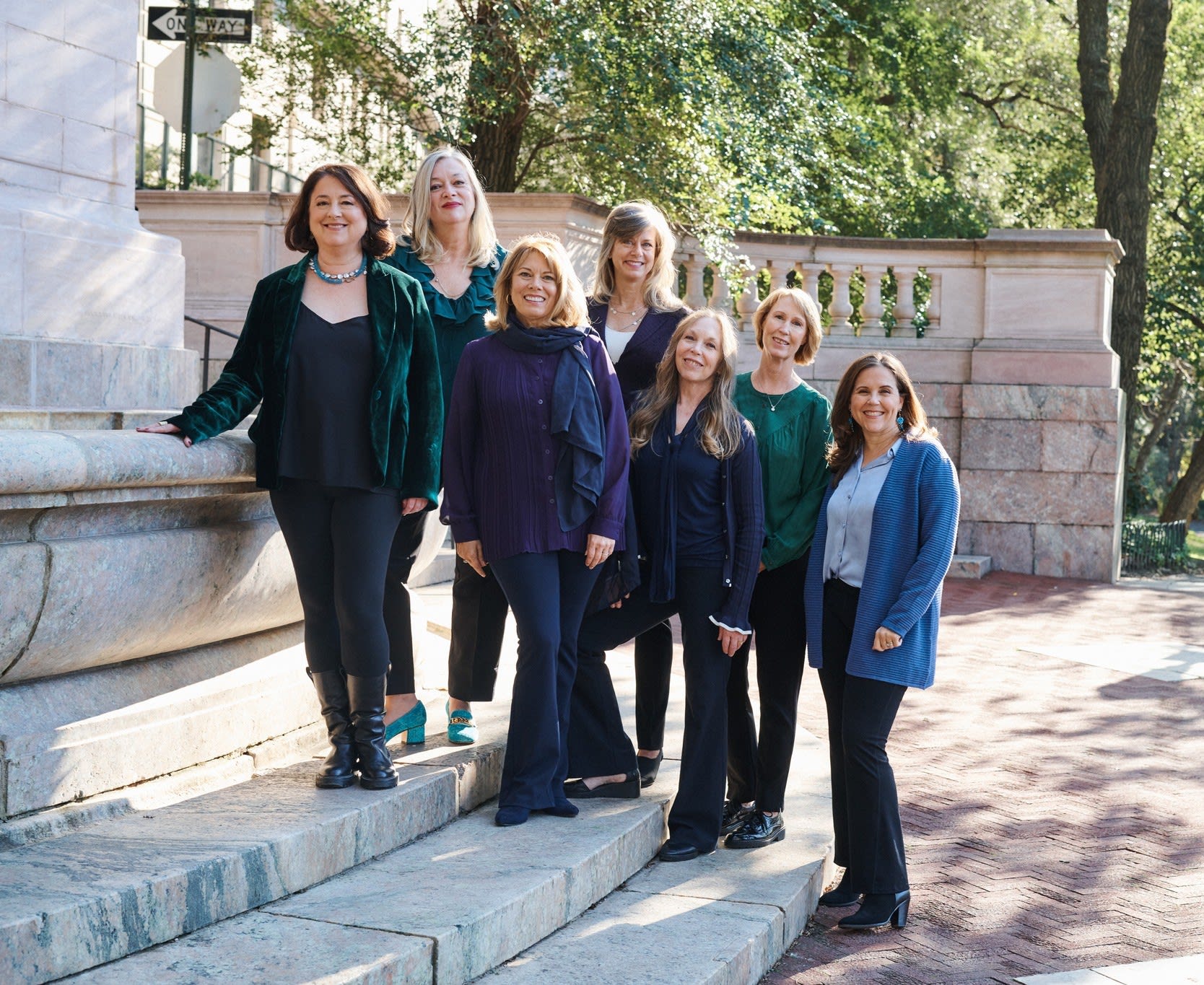 Seven women pose on stone steps beside a stone balustrade and leafy trees.