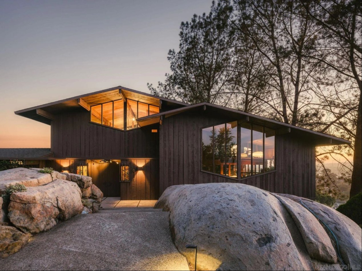 Dark wood and glass Engstrand Residence at 9450 Sunset Ave, La Mesa, lit at dusk behind large granite boulders