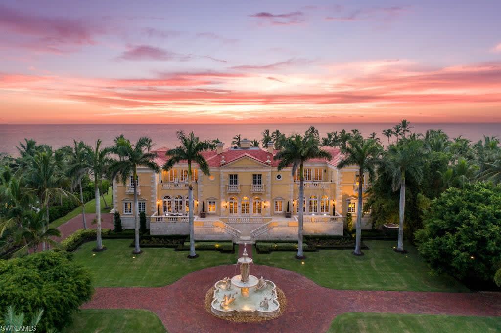Coastal mansion with palm trees and a fountain overlooking the ocean at sunset.