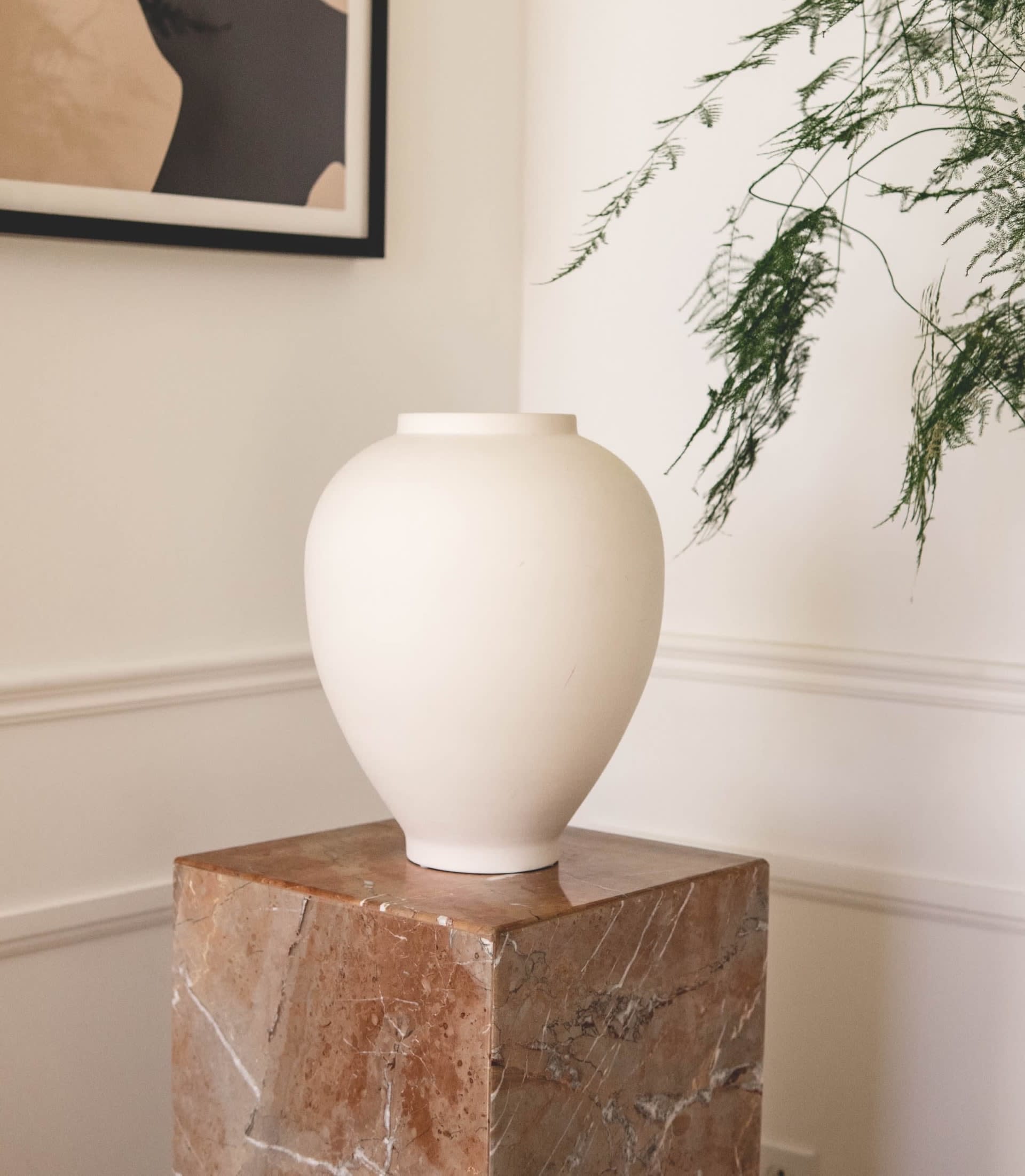 White ceramic vase on a reddish marble pedestal beneath framed abstract art and leafy branches.