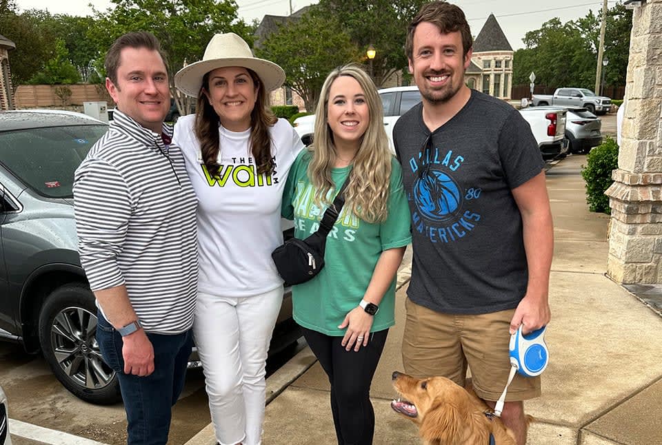 Men, women, and a dog posing for a photo on the sidewalk with trees and cars in the background