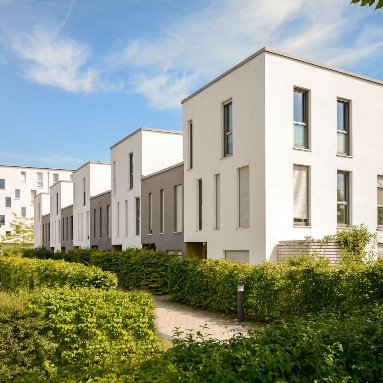 A row of modern, prefabricated white buildings with large windows, flat roofs, a front pathway, and surrounded by bushes.