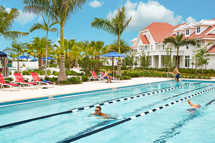 Outdoor lap pool with lane lines, palm trees, and a white building with red tile roofs.