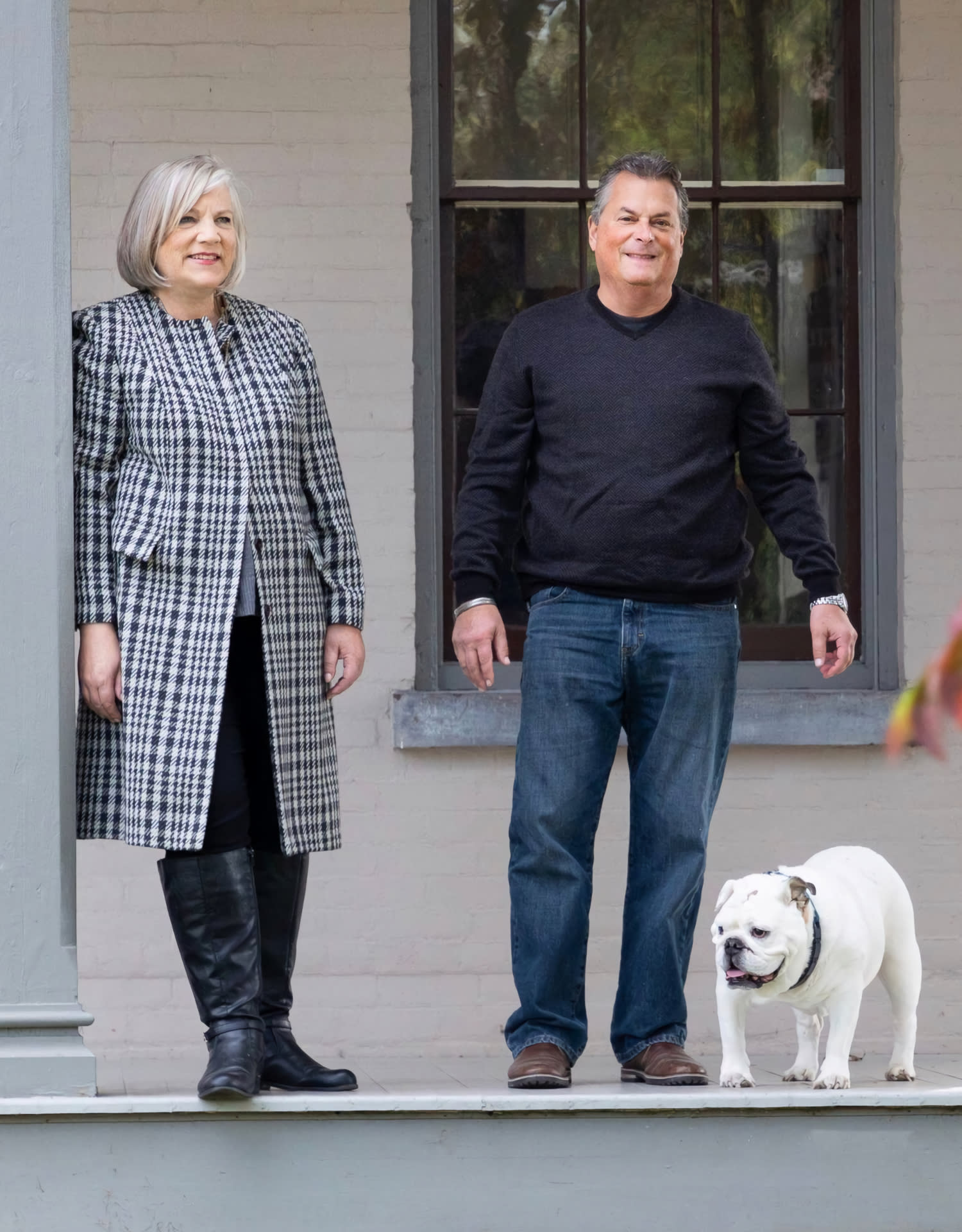 Two adults pose outside a house with a white bulldog.