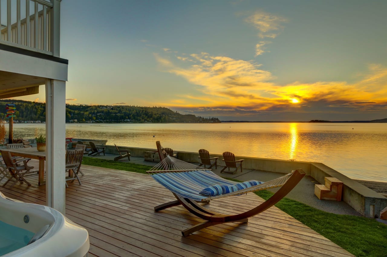 Lakeside deck with a hammock, Adirondack chairs, and hot tub overlooking a serene sunset over the water, surrounded by trees.