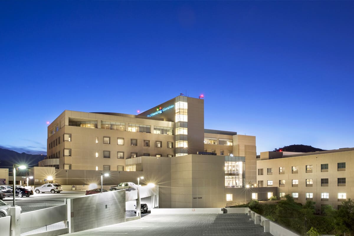 Glendale hospital with a beige facade, large windows, illuminated at night, parking in front, surrounded by hilly greenery.