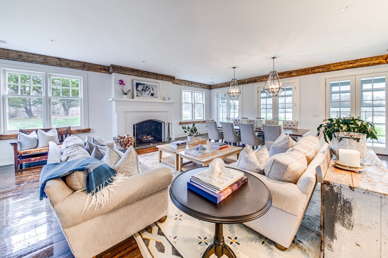 A living room with a light couch, rug, wooden armchair by the fireplace, framed picture, two windows, and hardwood floor.