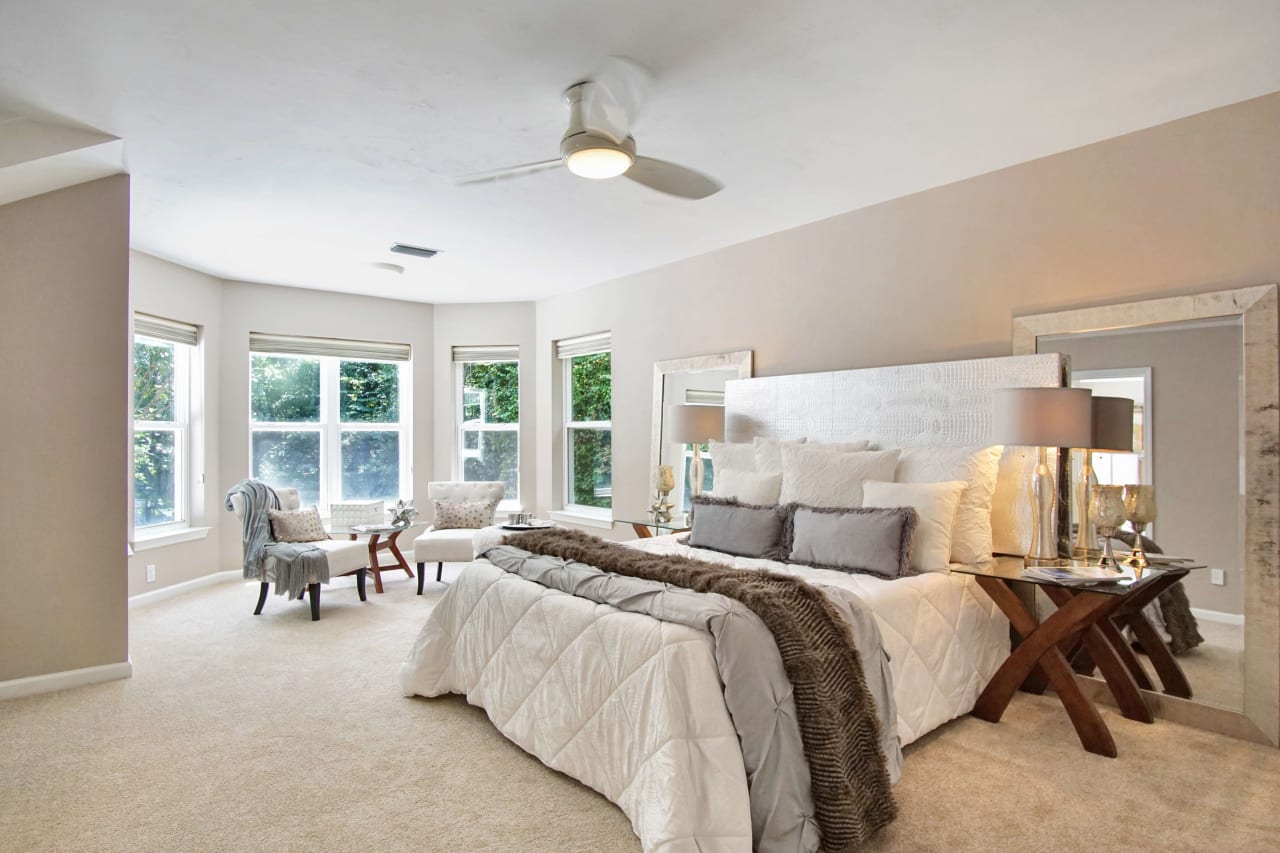 An interior view of a spacious bedroom with large windows, carpeted floors, and a ceiling fan. The room is furnished with a comfortable bed and seating area.