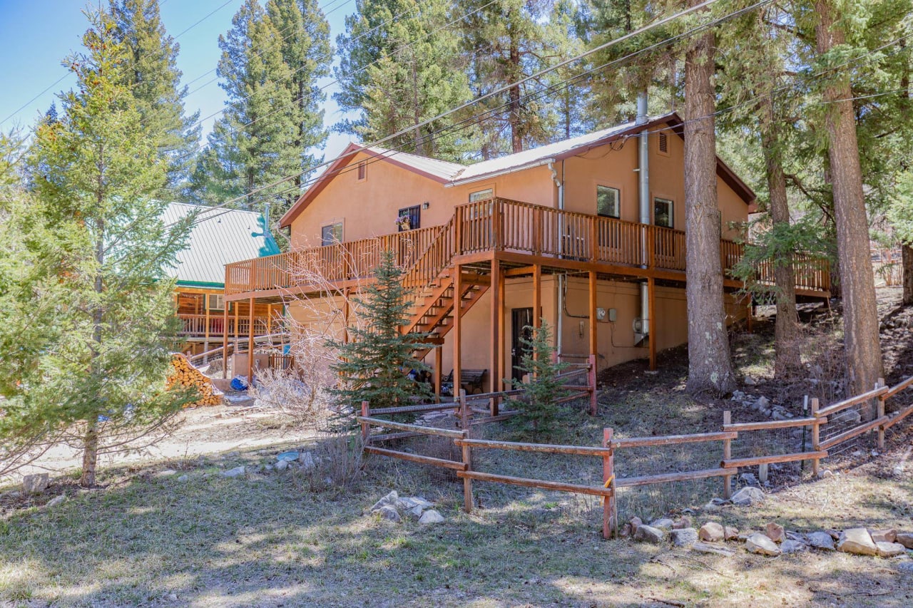 A wooden house in the middle of a forest. The house has a large deck, windows with shutters, and a chimney.