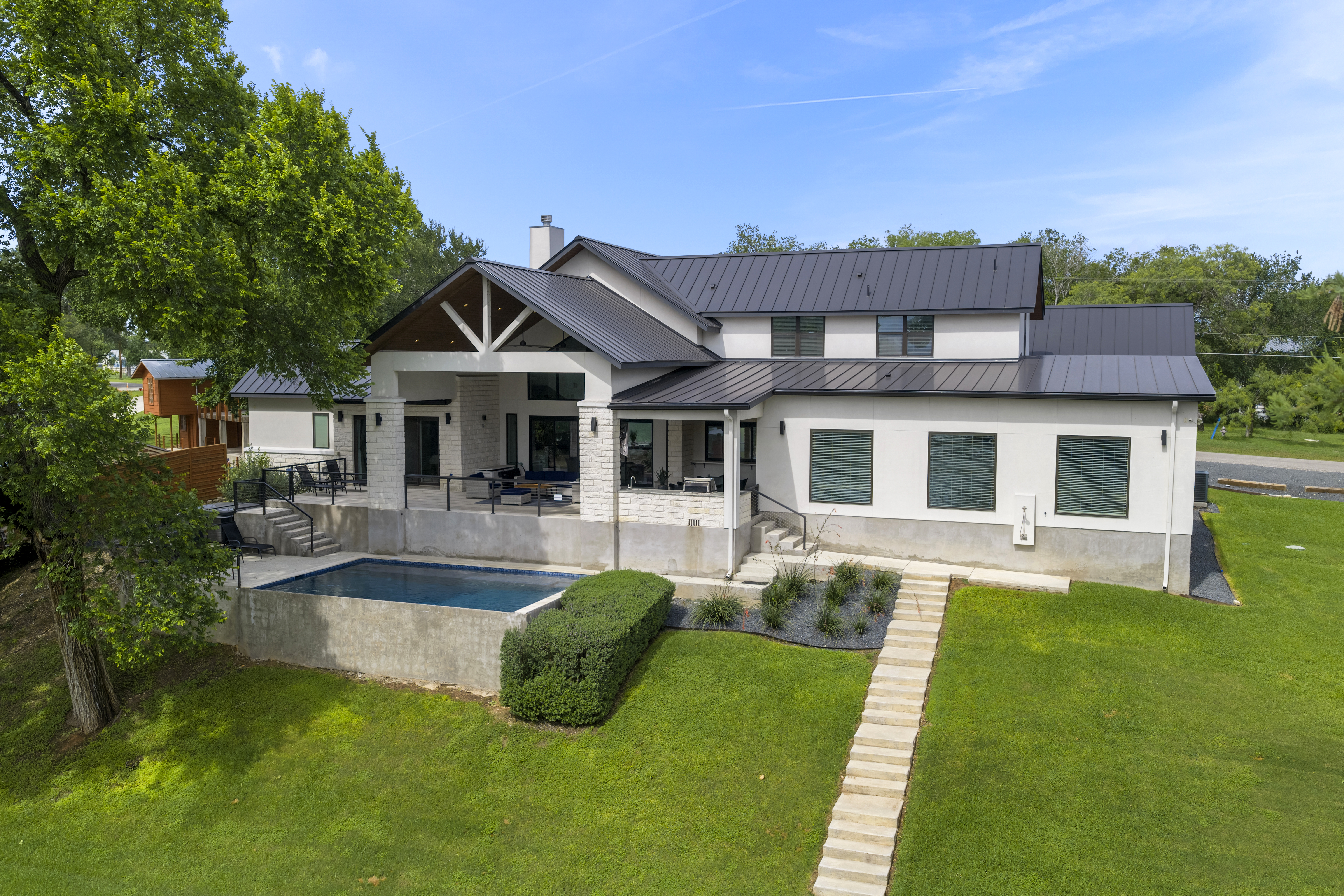 Rear of a two-story white home with a dark metal roof, a covered patio, an infinity-edge pool and a stone stairway down a green lawn
