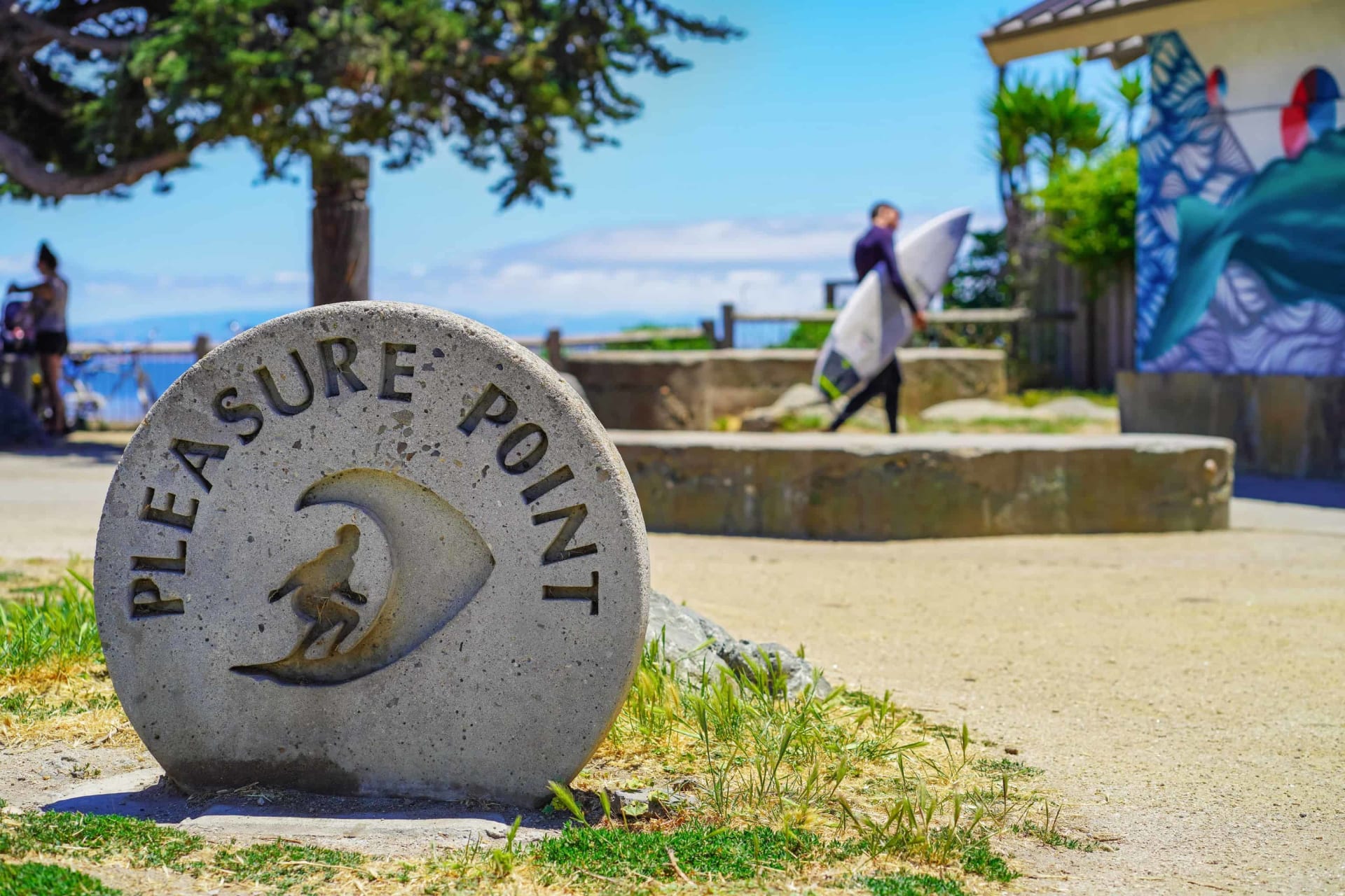 Pleasure Point marker beside a coastal path as surfers carry boards near the ocean.