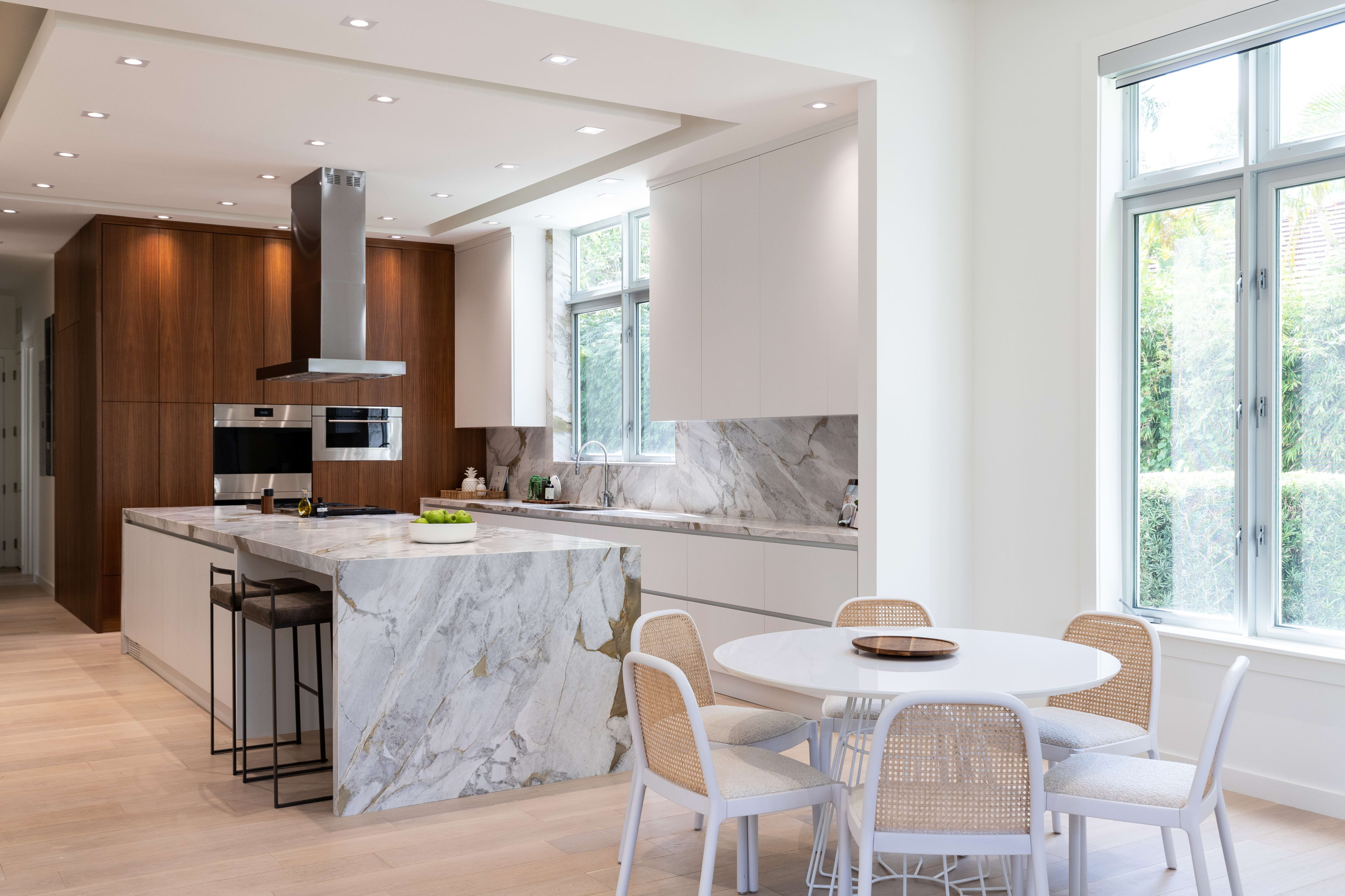 A kitchen with a marble island, dark wood cabinets, stainless steel appliances, a dining table, and outside-view windows.