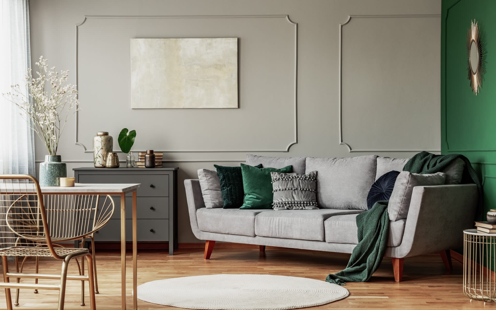 A living room with a gray couch, green pillows, a round rug, a painting on the wall, and large windows.