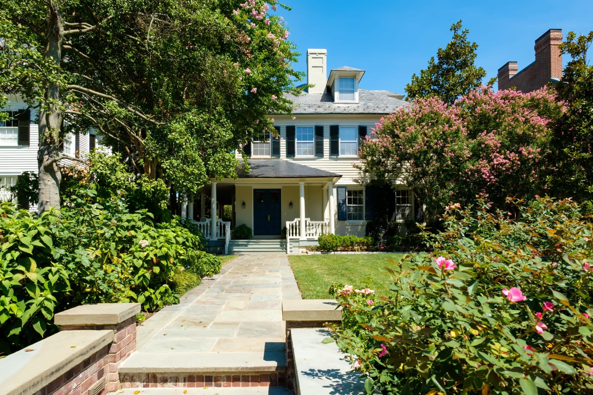 A white house with a brick walkway to the front door, dormer windows, and surrounded by mature trees and flowering shrubs.