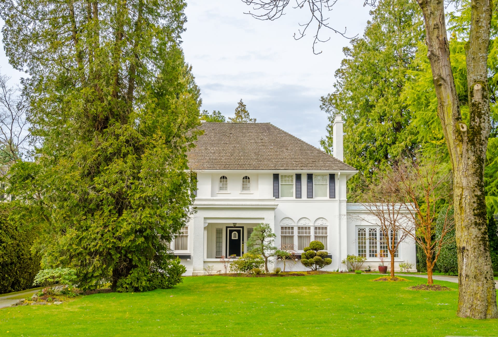 A large white house with a slate roof and a chimney, a long driveway to the front door, a lush lawn, and surrounded by trees.