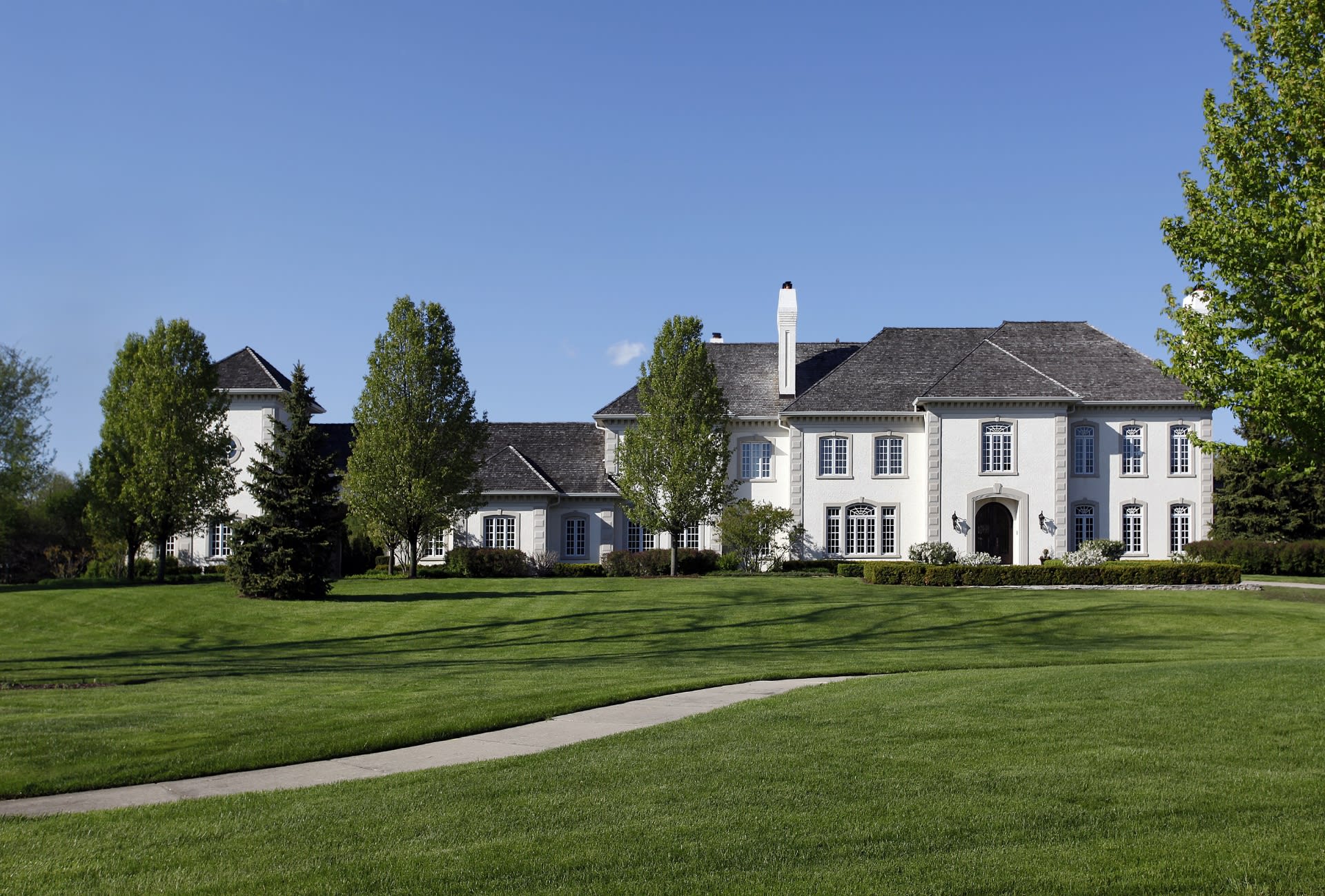 A large white house with a slate roof, chimney, and long gravel driveway sits on a green lawn, surrounded by scattered trees.